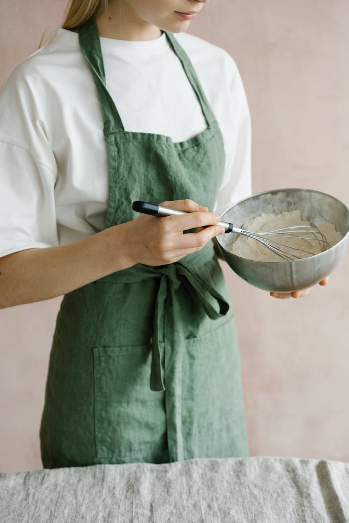 Woman in green apron whisking mixture in a steel bowl, illustrating home cooking.