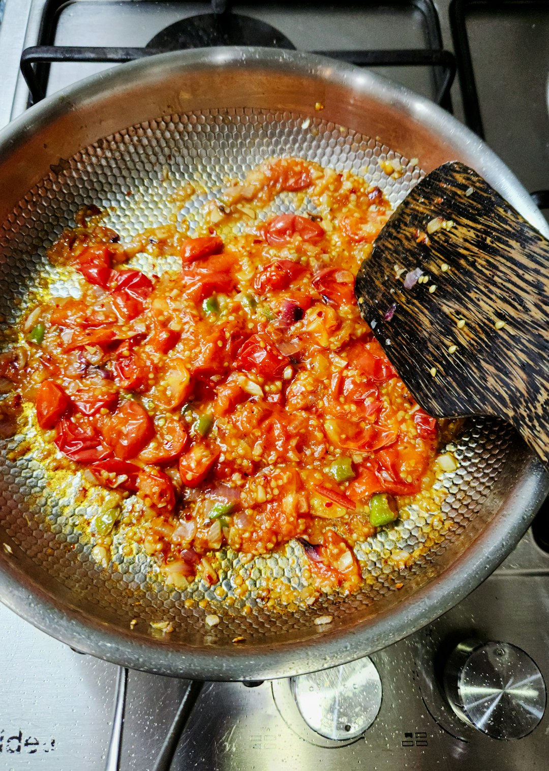about-02 Ingredients being sautéed in a textured stainless steel pan on a stovetop. Chopped tomatoes, onions, and green chilies are simmering in oil, glistening with spices. This mixture likely forms the aromatic base (masala) for a Sri Lankan or South Asian curry or dish.